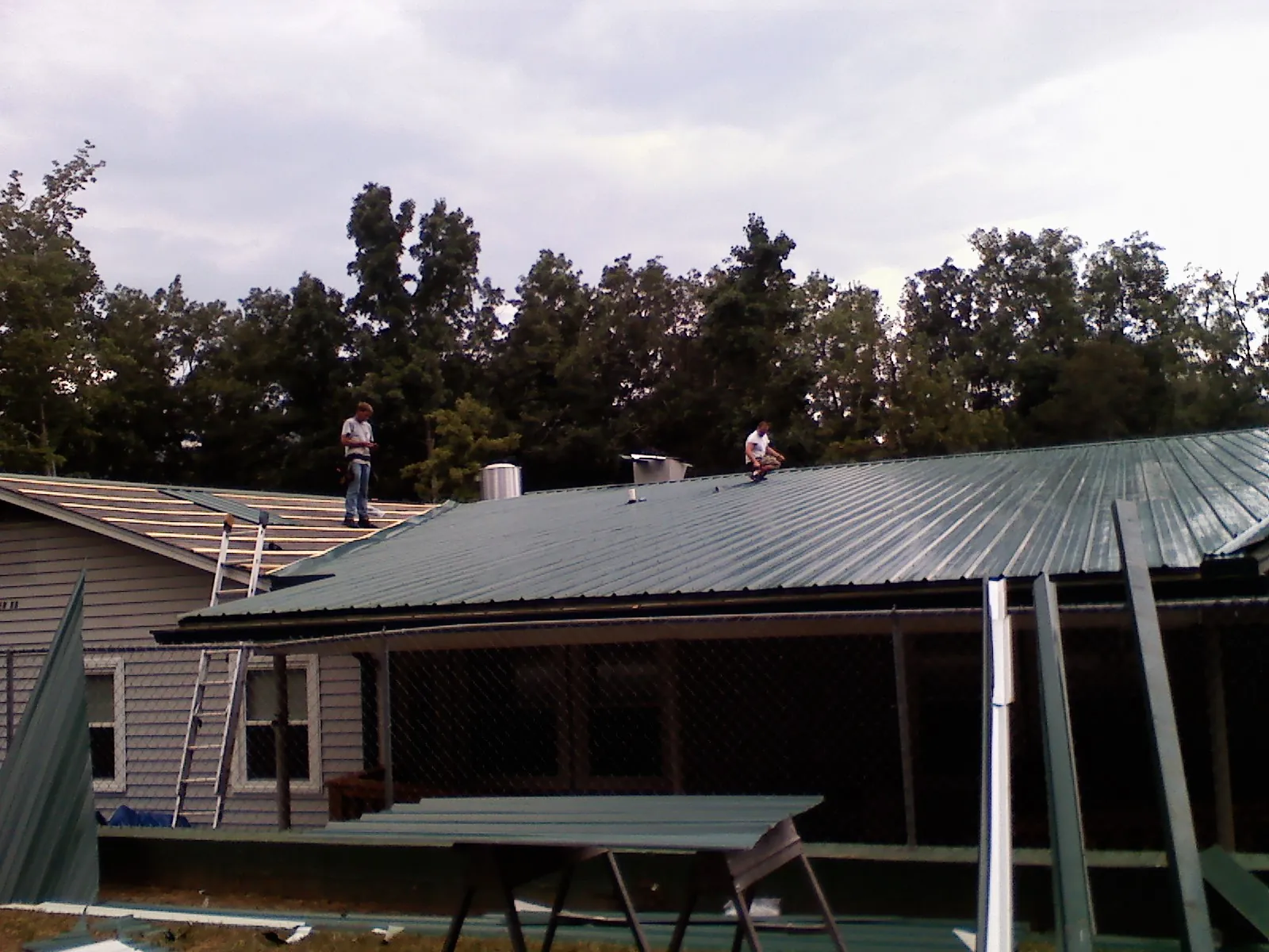 Two roofers installing green metal panels above a covered porch in Sugar Grove